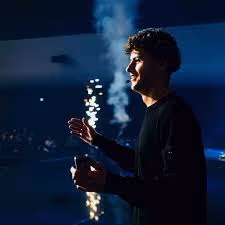 Man in dark clothing holding a lit sparkler indoors with smoke rising in a dimly lit room.