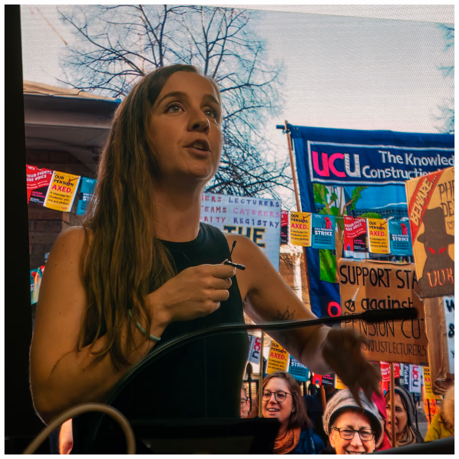 Woman speaking passionately at a protest rally with banners and signs supporting staff and opposing pension cuts.