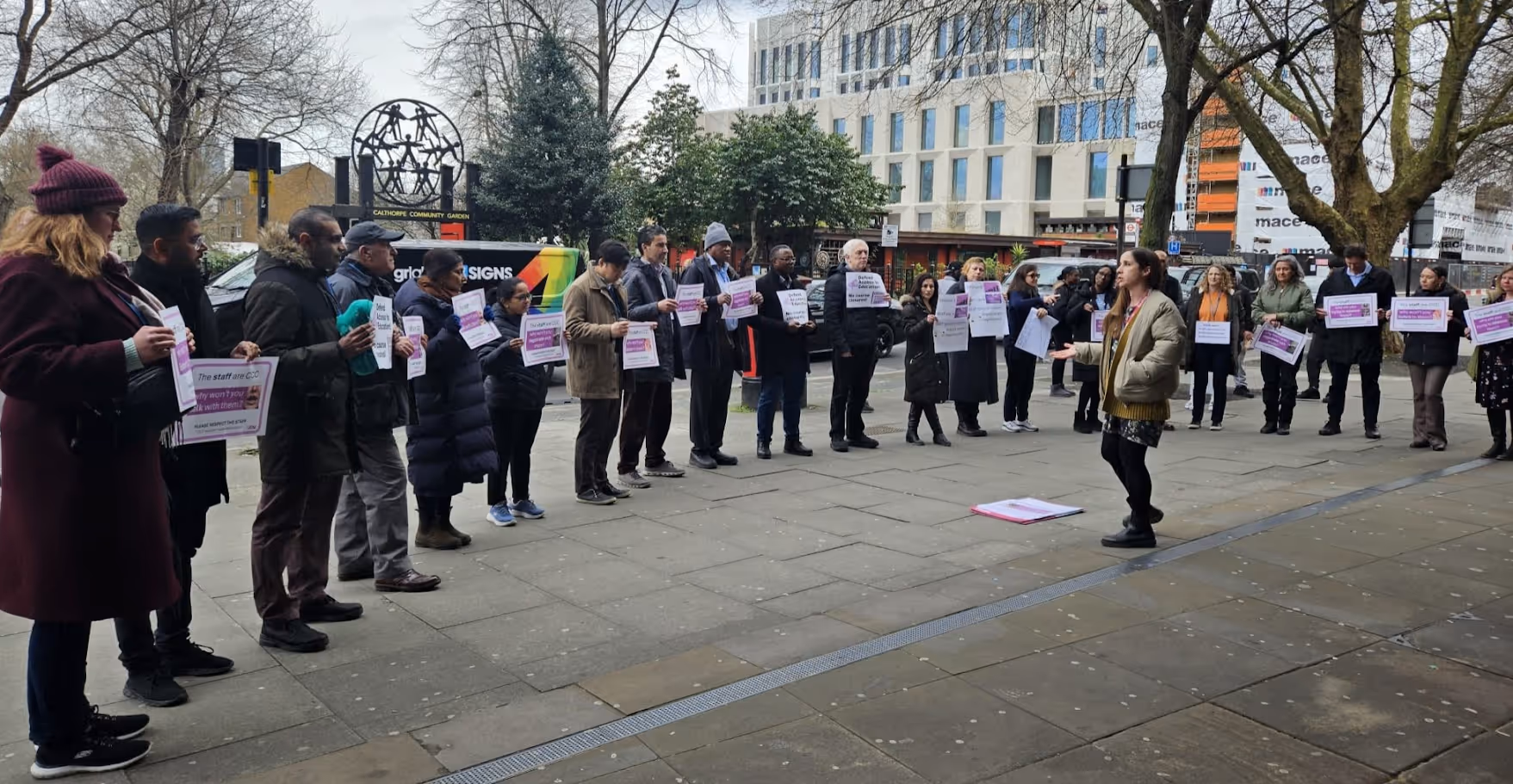 A group of people standing outdoors in a semi-circle holding protest signs, with a woman speaking to them on a city sidewalk.