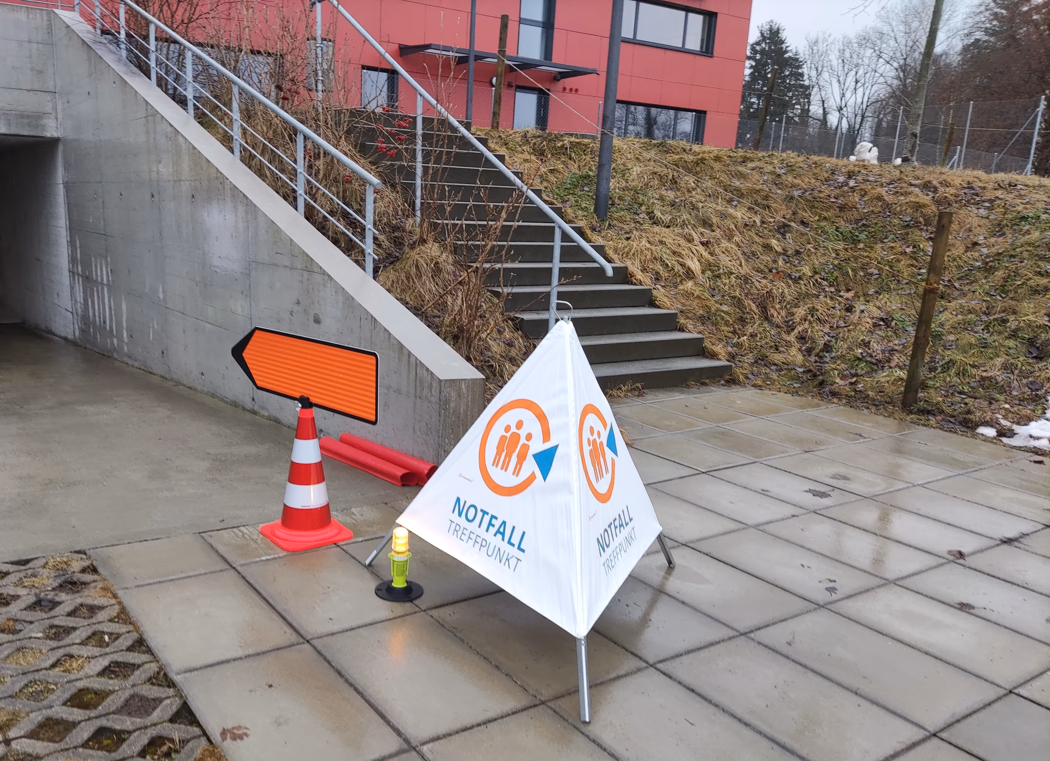 Notfalltreffpunkt-Signal mit weißem Dreieck und orangem Symbol neben einem orangenes Richtungsschild auf einer nassen Betonfläche vor einer Treppe und einem roten Gebäude.