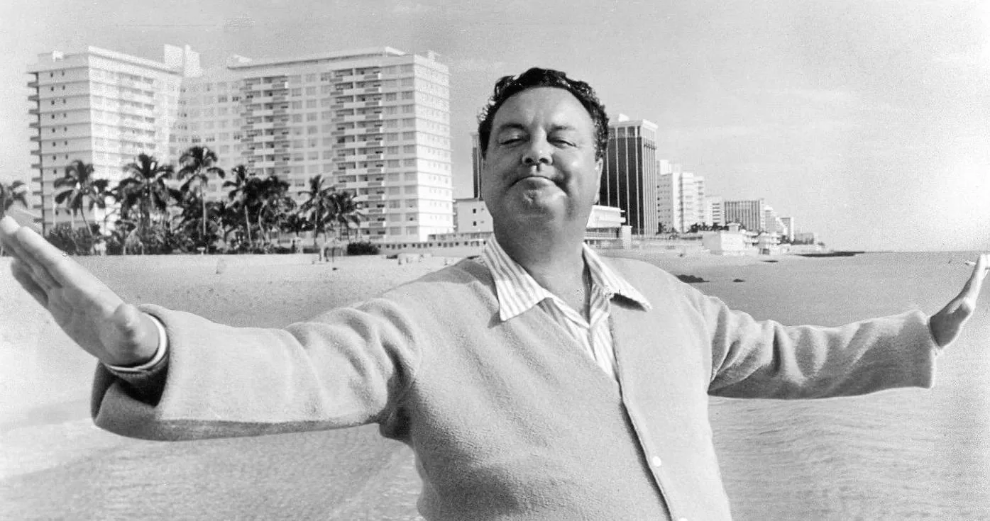 Black and white photo of a man standing on a beach with arms outstretched, smiling with buildings and palm trees in the background.