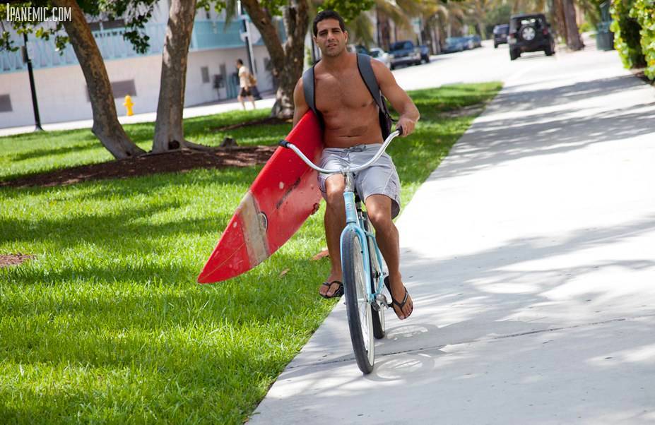 Shirtless man in shorts and flip-flops riding a bicycle on a sidewalk while carrying a red surfboard under his arm.