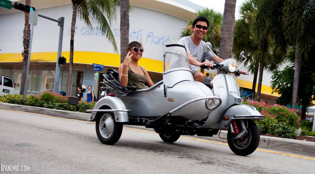 Two people wearing sunglasses riding a silver scooter with a sidecar on a city street lined with palm trees.