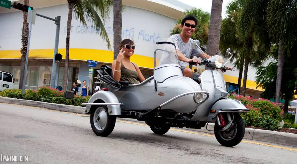 Two people wearing sunglasses riding a silver scooter with a sidecar on a city street lined with palm trees.