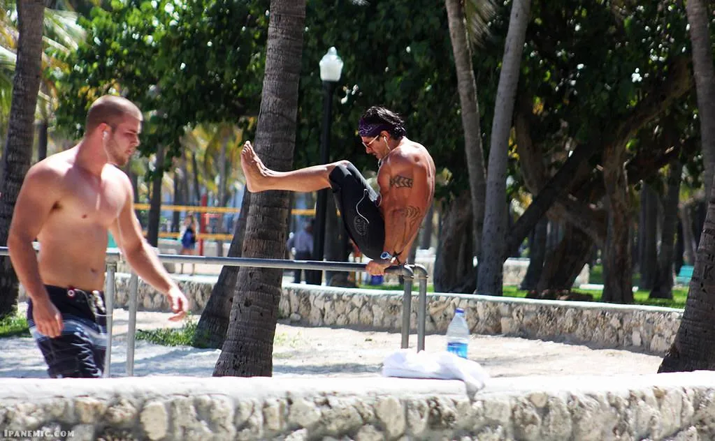 Shirtless man performing a workout L-sit on parallel bars outdoors with another shirtless man walking nearby.