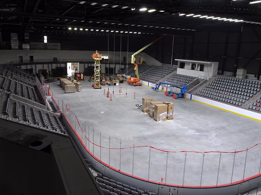 Indoor ice hockey rink under construction with empty seats, lifts, and equipment on the concrete floor.