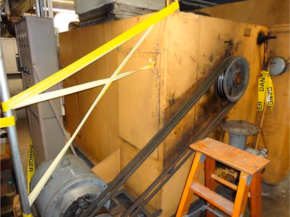 Industrial machinery with belts and pulleys beside a wooden step stool, marked with yellow caution tape.