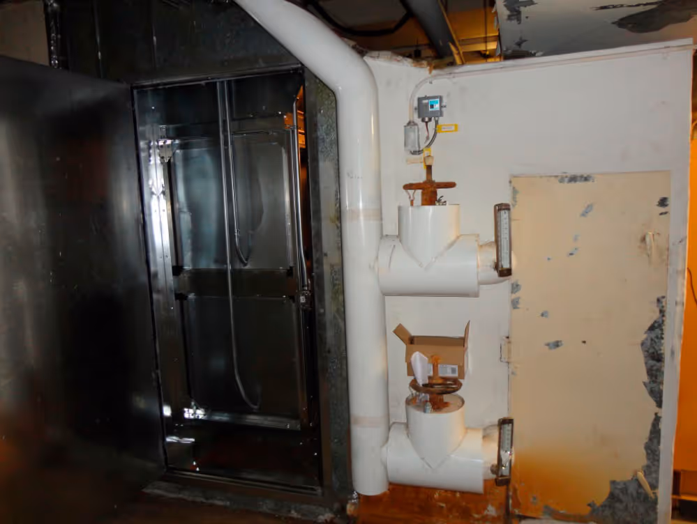 Industrial pipes and valves next to a metal panel and a worn, rusty door in a dimly lit room.