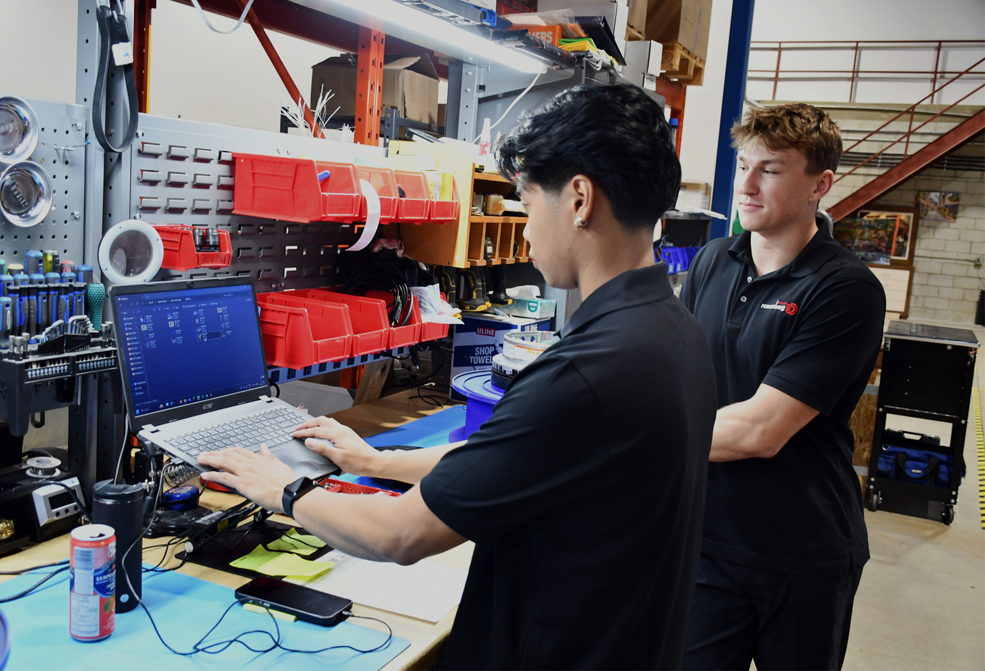Two people working at a workshop bench with a laptop and organized tools on the wall.