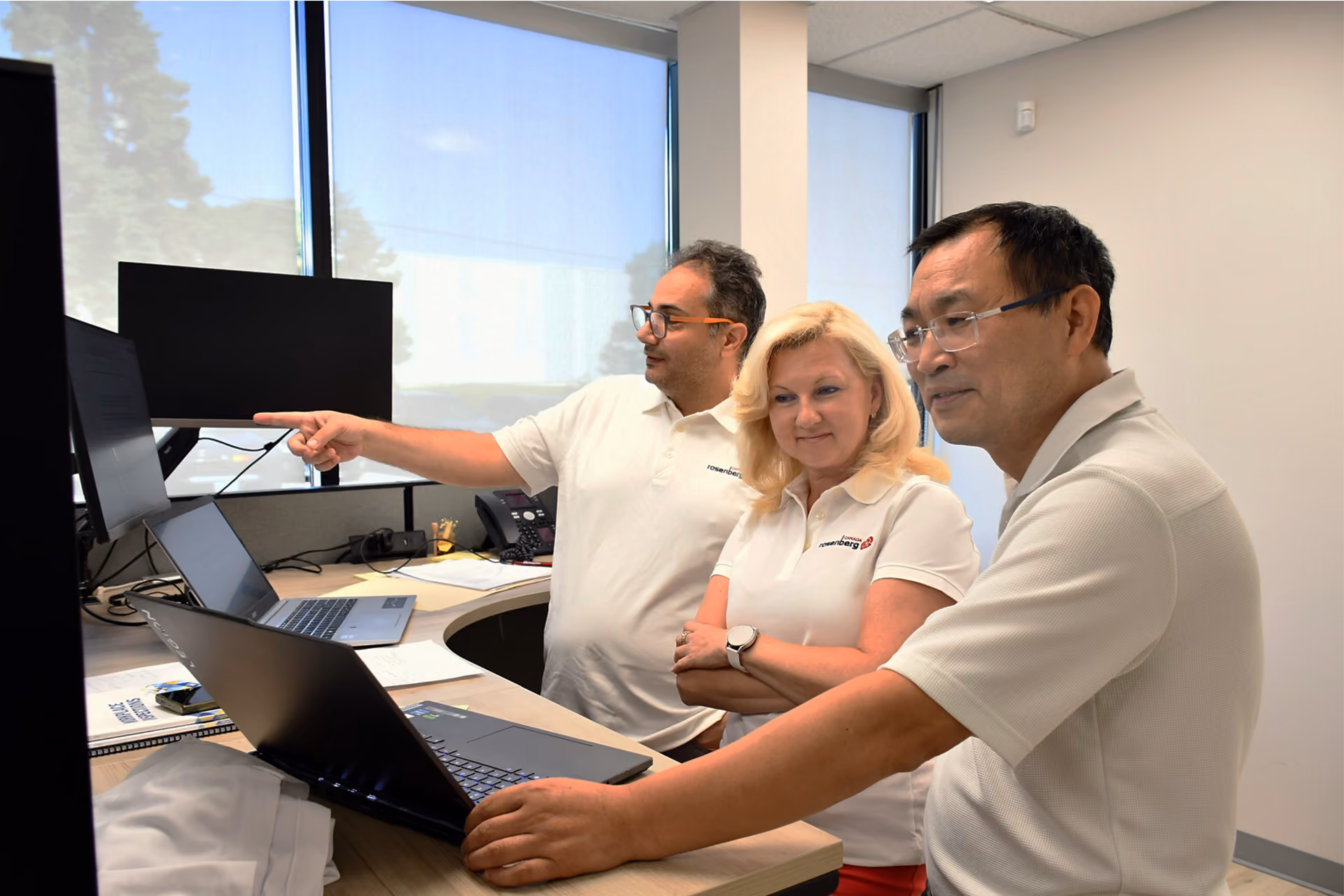 Three colleagues in matching white polos collaborating at a curved desk with laptops and monitors.