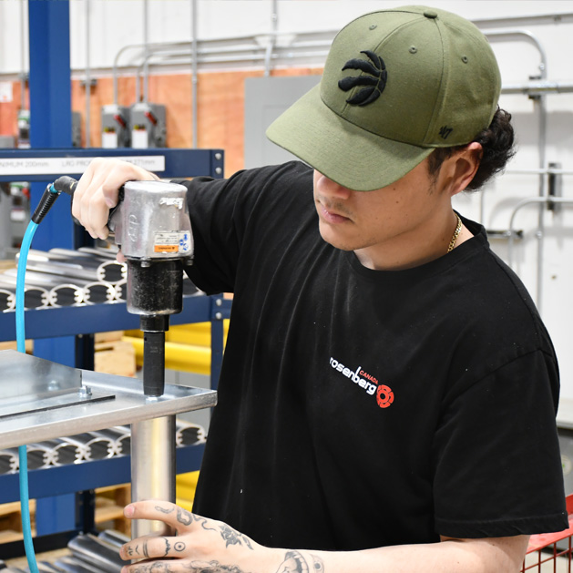Man wearing a green cap and black shirt uses a tool to fasten a metal piece in a workshop.