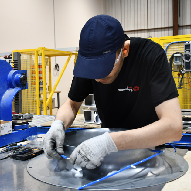 Worker in gloves and cap assembling or inspecting metal parts in an industrial workshop.