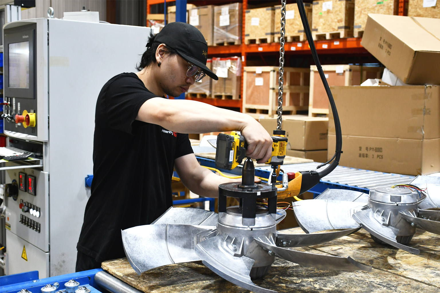 Technician in black shirt and cap assembling metal fan blades using a drill in a factory warehouse.