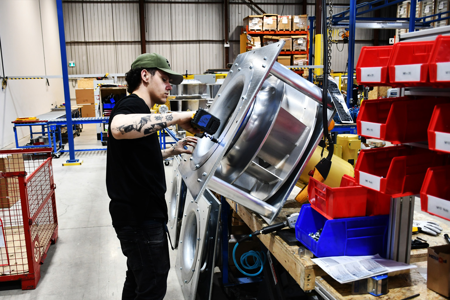 Worker with tattoos using drill on large metal industrial component in a factory setting.