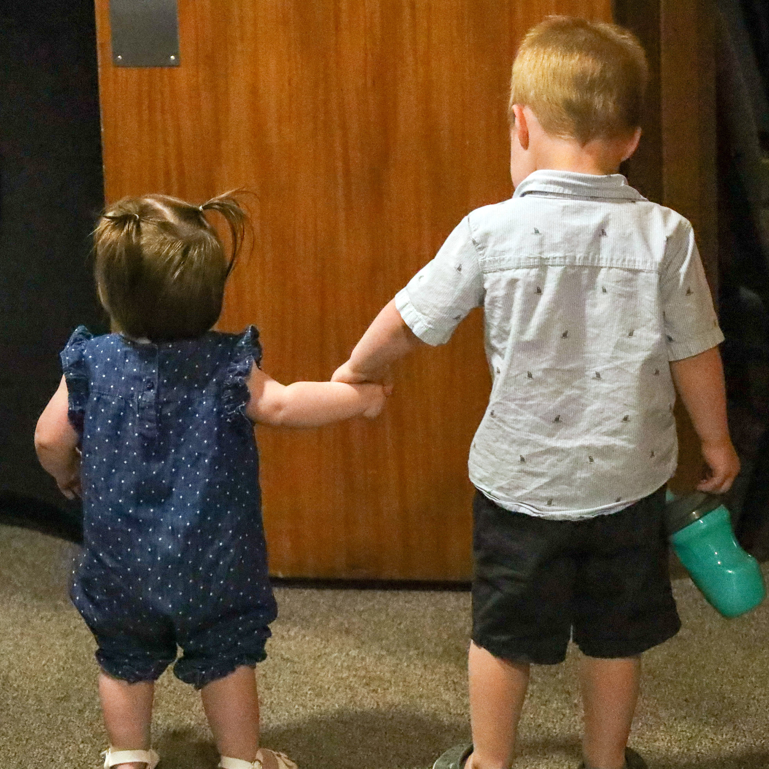 Toddler boy holding hands with a younger toddler girl inside a room, both seen from behind.
