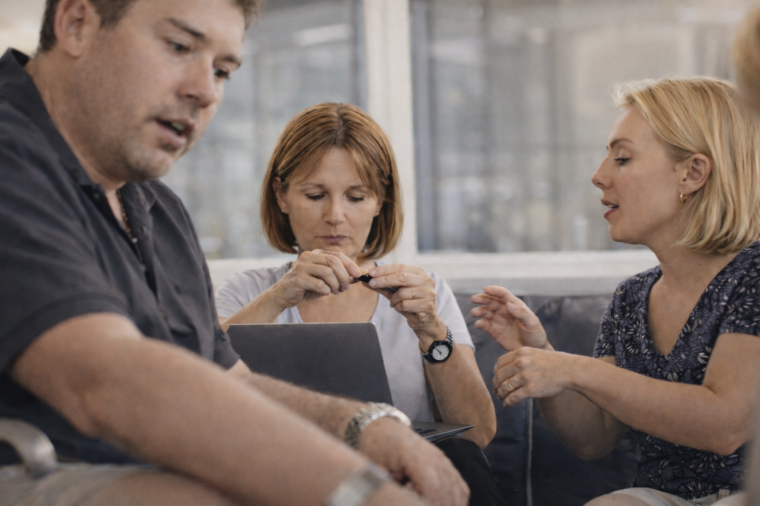 Three adults seated indoors, one woman in the center holding a small object with a laptop in front of her, a man in the foreground and another woman on the right gesturing with her hands.