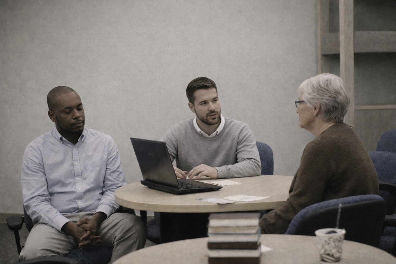 Three people sitting around a table engaged in conversation with a laptop and documents in front of them.