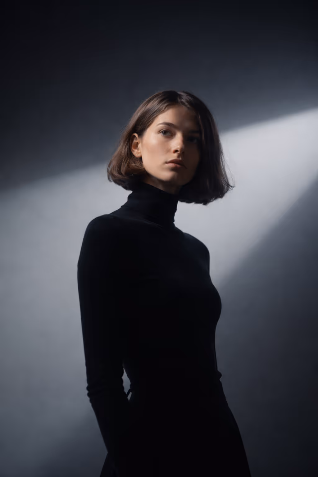 Woman with short brown hair wearing a black turtleneck, illuminated by dramatic side lighting against a dark background.