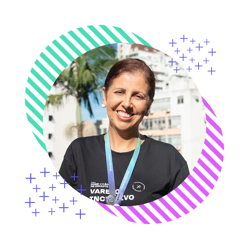 A diretora Carla Inácio sorridente, com cabelos escuros presos, vestindo uma camisa preta e um cordão azul, em pé ao ar livre com prédios e uma palmeira ao fundo.