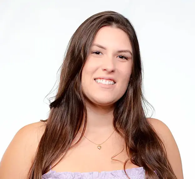 Daniela Silva sorridente com longos cabelos castanhos, usando uma blusa lilás clara contra um fundo branco.