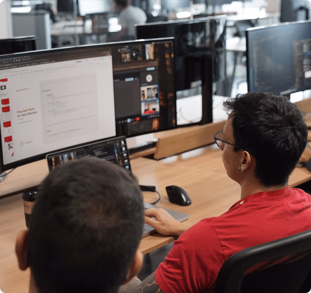 Two men collaborating at a desk with multiple computer screens displaying documents and videos in an open office.