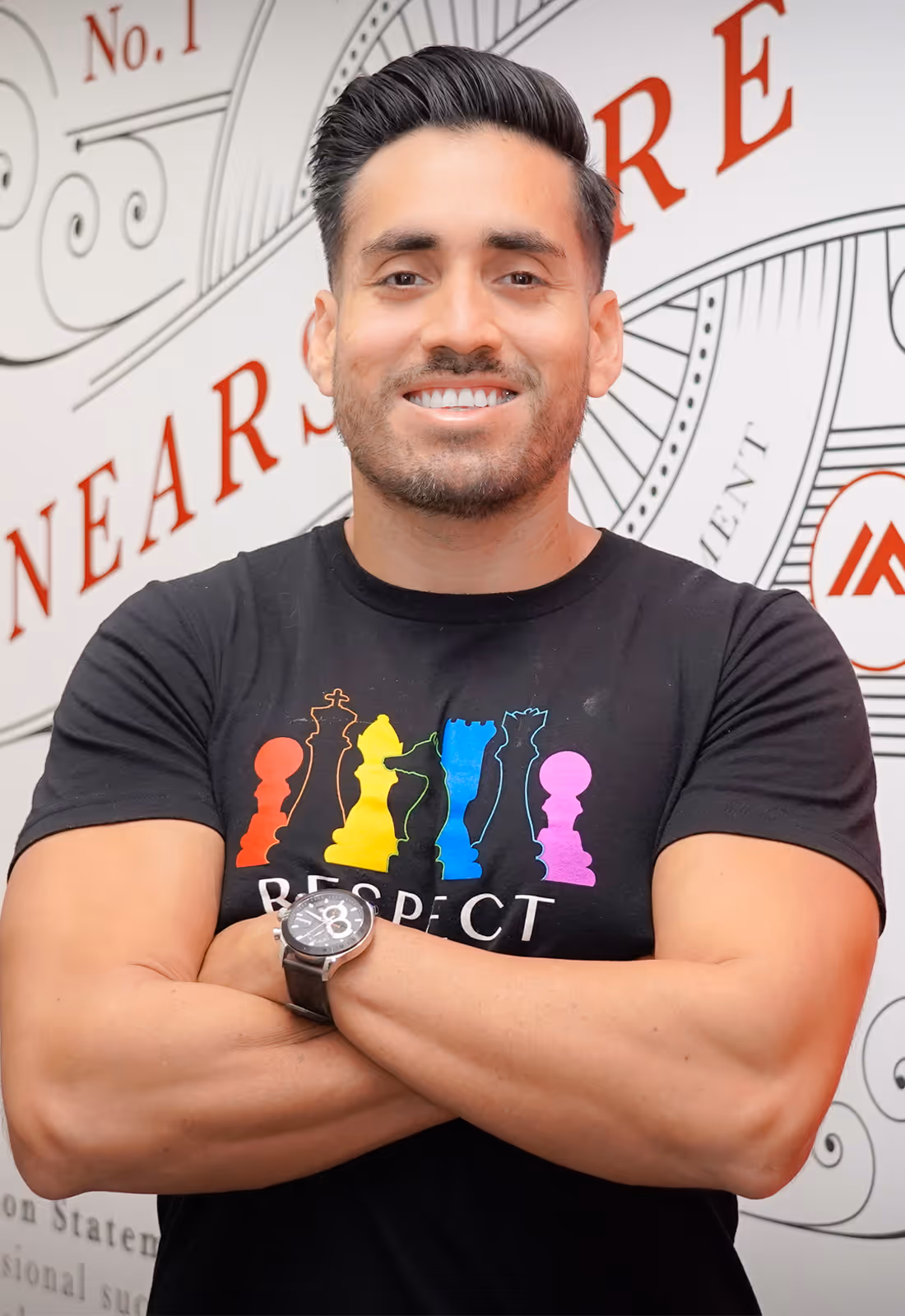 Man smiling with crossed arms wearing a black t-shirt featuring colorful chess pieces and the word RESPECT.
