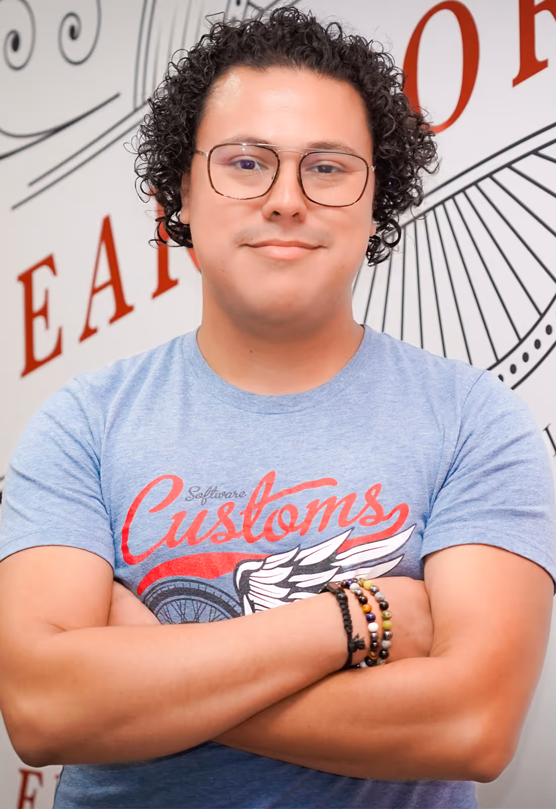 Man with curly hair and glasses wearing a blue t-shirt with the word 'Customs' and a winged wheel graphic, standing with arms crossed.