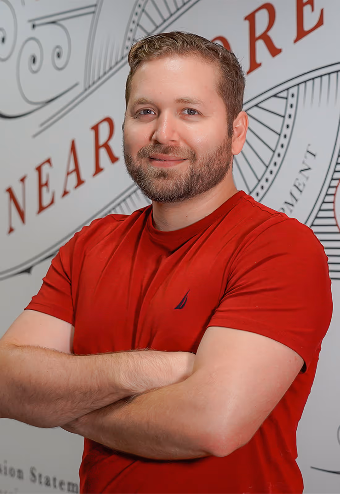 Smiling man with light brown hair and beard wearing a red t-shirt with crossed arms in front of a decorative wall with text.