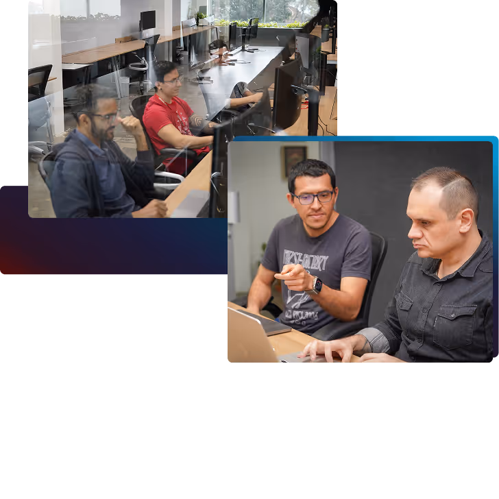 Two men working collaboratively at a desk with a laptop, while in the background two other men are seated at computers in an office environment.