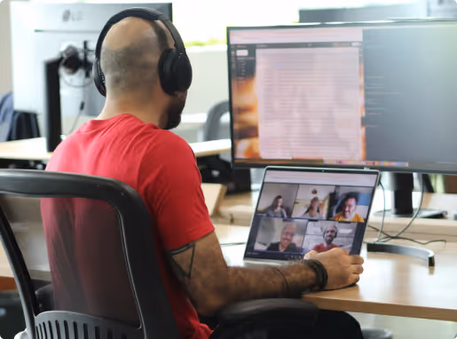 Man wearing headphones and red shirt in an office participating in a video conference on a laptop with six people visible on the screen.