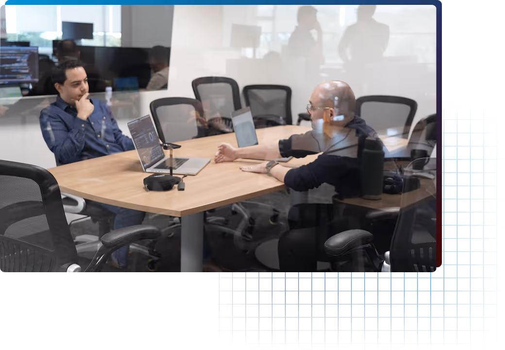 Two men in an office meeting room engaged in a discussion with laptops open on the table.