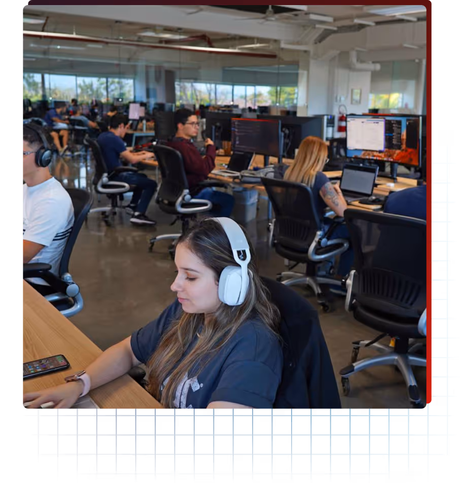 Office space with multiple people working at desks with laptops and monitors, one woman in foreground wearing headphones.