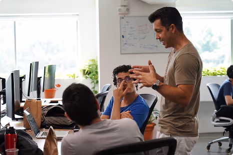 Two men having an animated discussion in an office with computers and a whiteboard in the background.