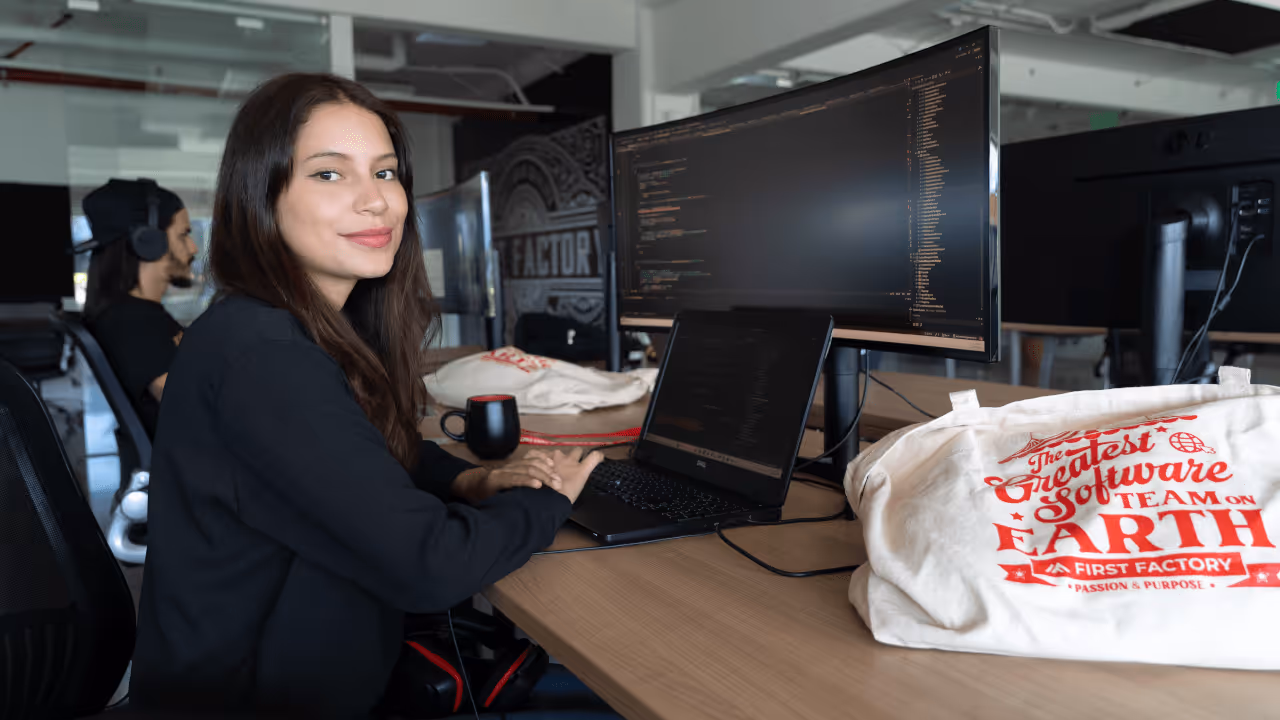 Young woman sitting at a desk smiling, working on a laptop connected to a large monitor displaying code, with a tote bag reading 'The Greatest Software Team on Earth' nearby.