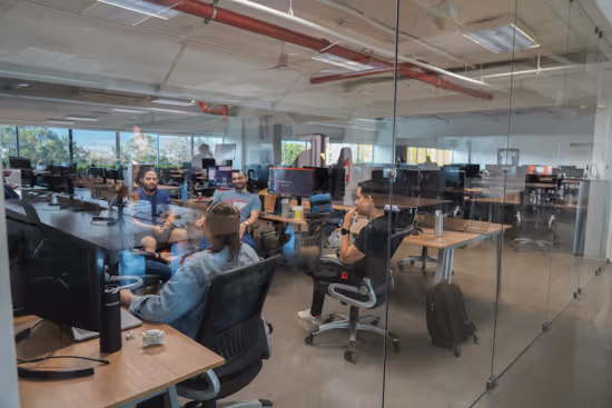Four colleagues having a discussion inside a modern glass-walled office with computers and desks.