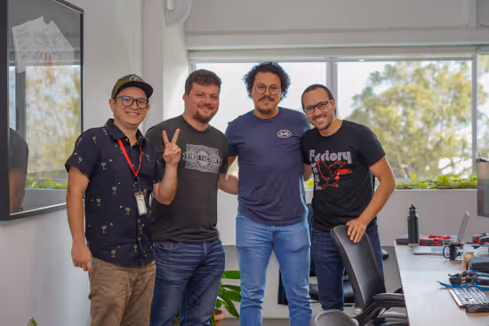 Group of four men standing together and smiling in a bright office setting with large windows and a desk with gadgets.