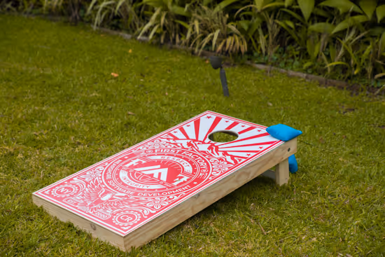 Red and white cornhole board with blue bean bags on grass in a garden.