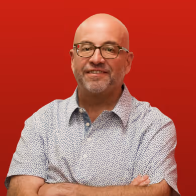 Smiling bald man with glasses wearing a patterned short-sleeve shirt against a red background.