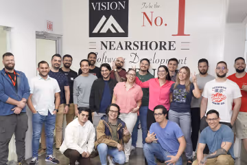 Group of diverse young adults smiling and posing together in an office hallway in front of a wall displaying the words Vision, Nearshore Software Development, and To be the No. 1.