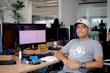 Man wearing a baseball cap and gray graphic t-shirt sitting at a desk with multiple computer monitors in an office.