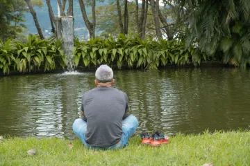 Man sitting cross-legged on grass facing a pond with fish and a small waterfall, with red shoes placed beside him.