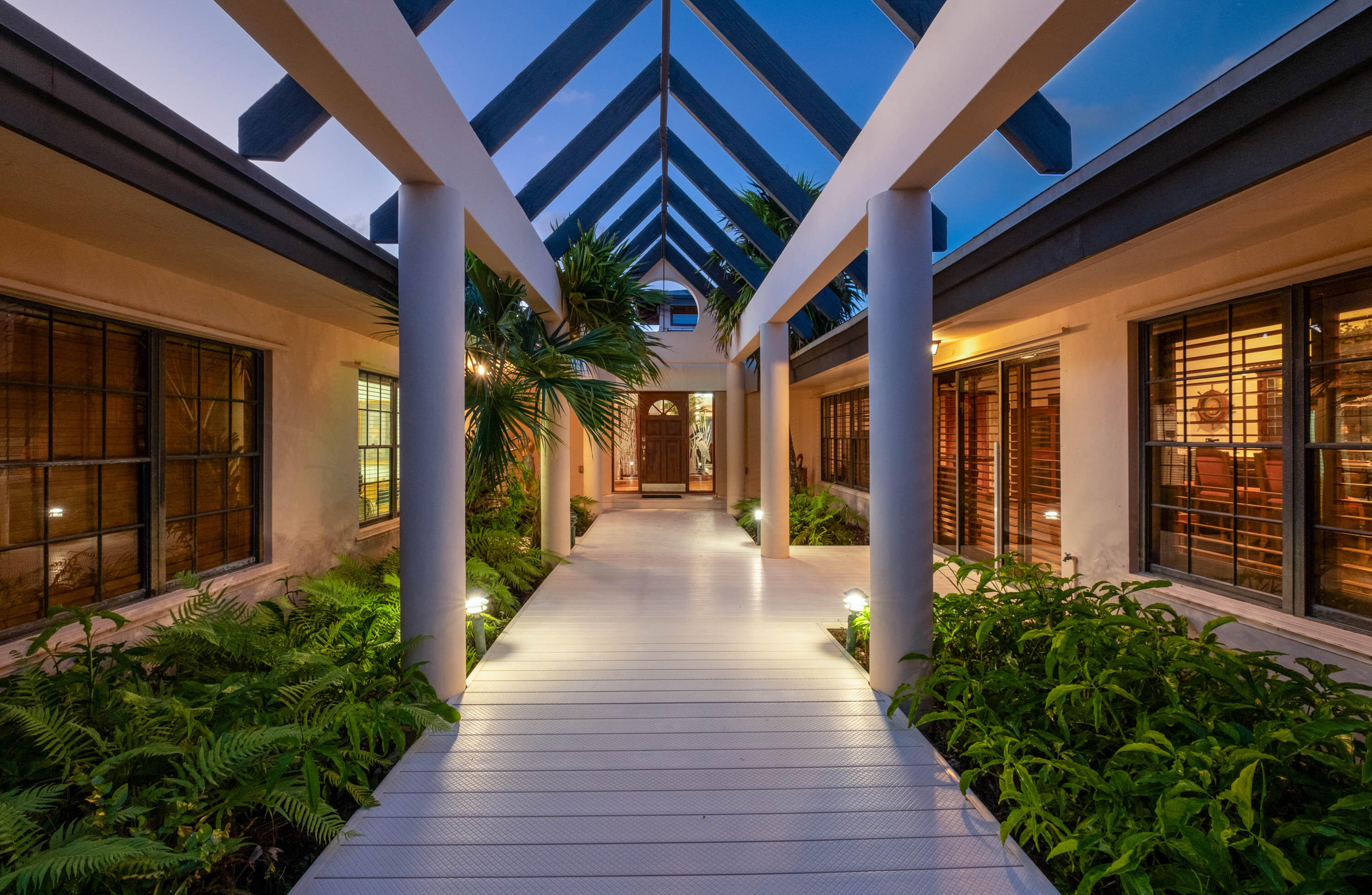 Well-lit walkway with white floor and columns, flanked by green plants, under a blue pitched roof leading to a wooden door at dusk.
