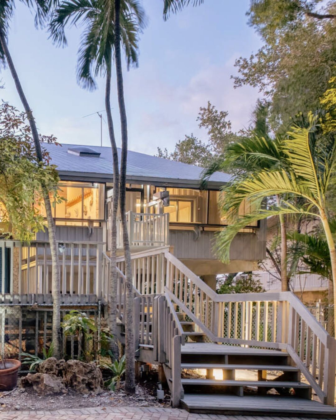 Elevated wooden house with stairs and railings surrounded by palm trees and greenery at dusk.