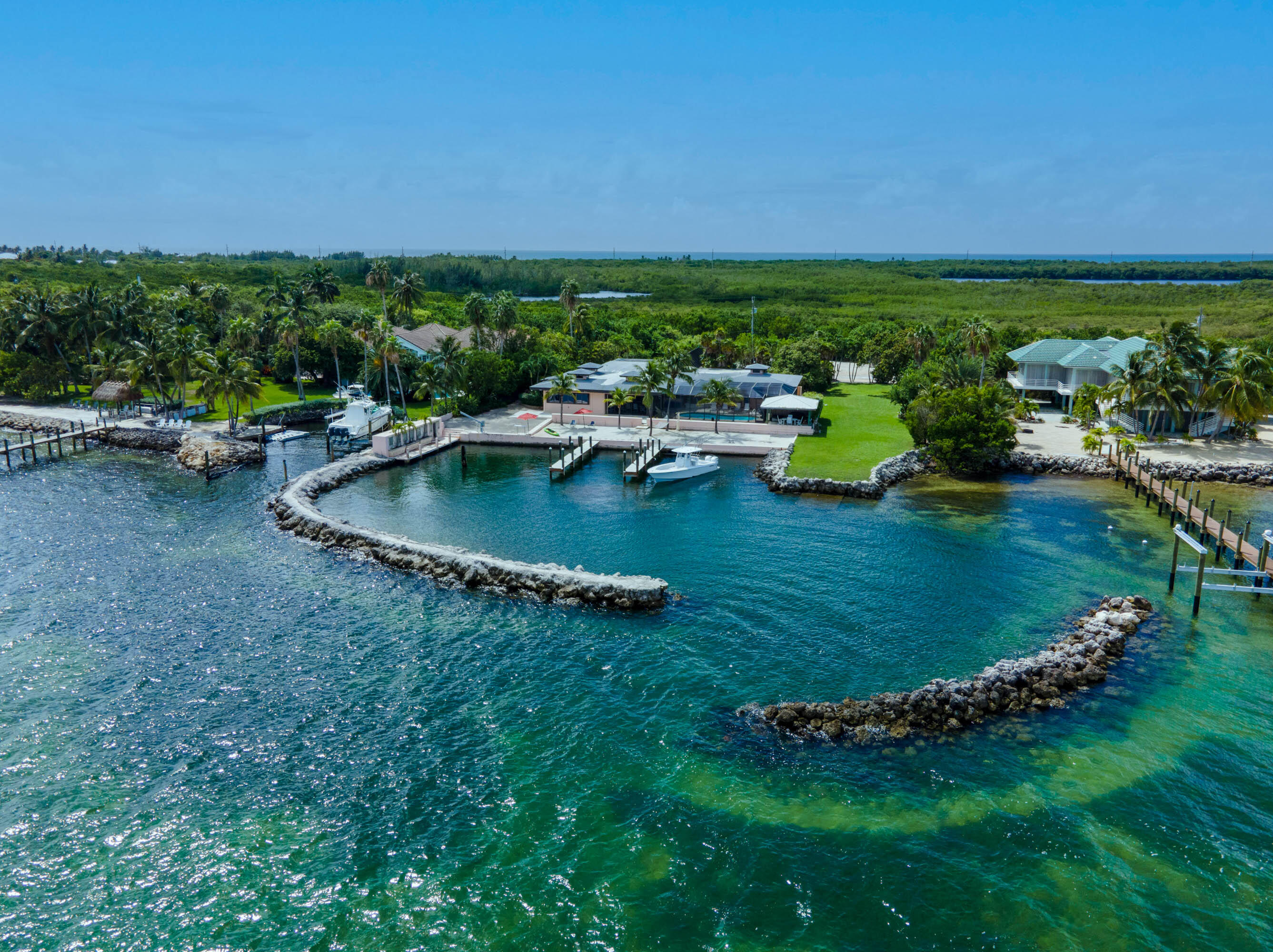 Aerial view of a coastal property with two houses, docks, boats, green lawns, palm trees, and a rocky seawall enclosing a calm blue-green bay.