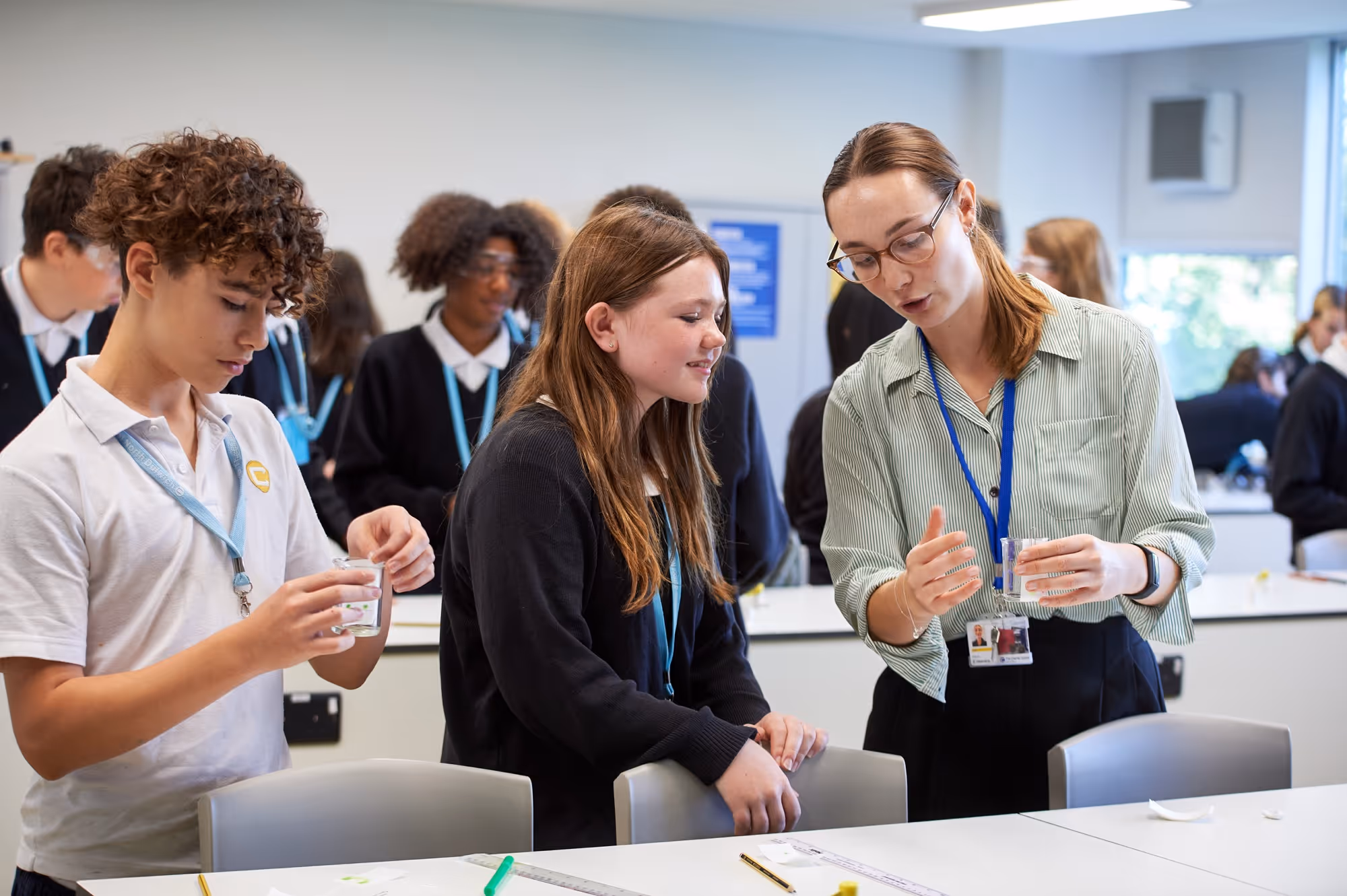 A teacher wearing glasses explains a science experiment to two students in a classroom; the teacher and one student hold clear containers while others engage in activities in the background.