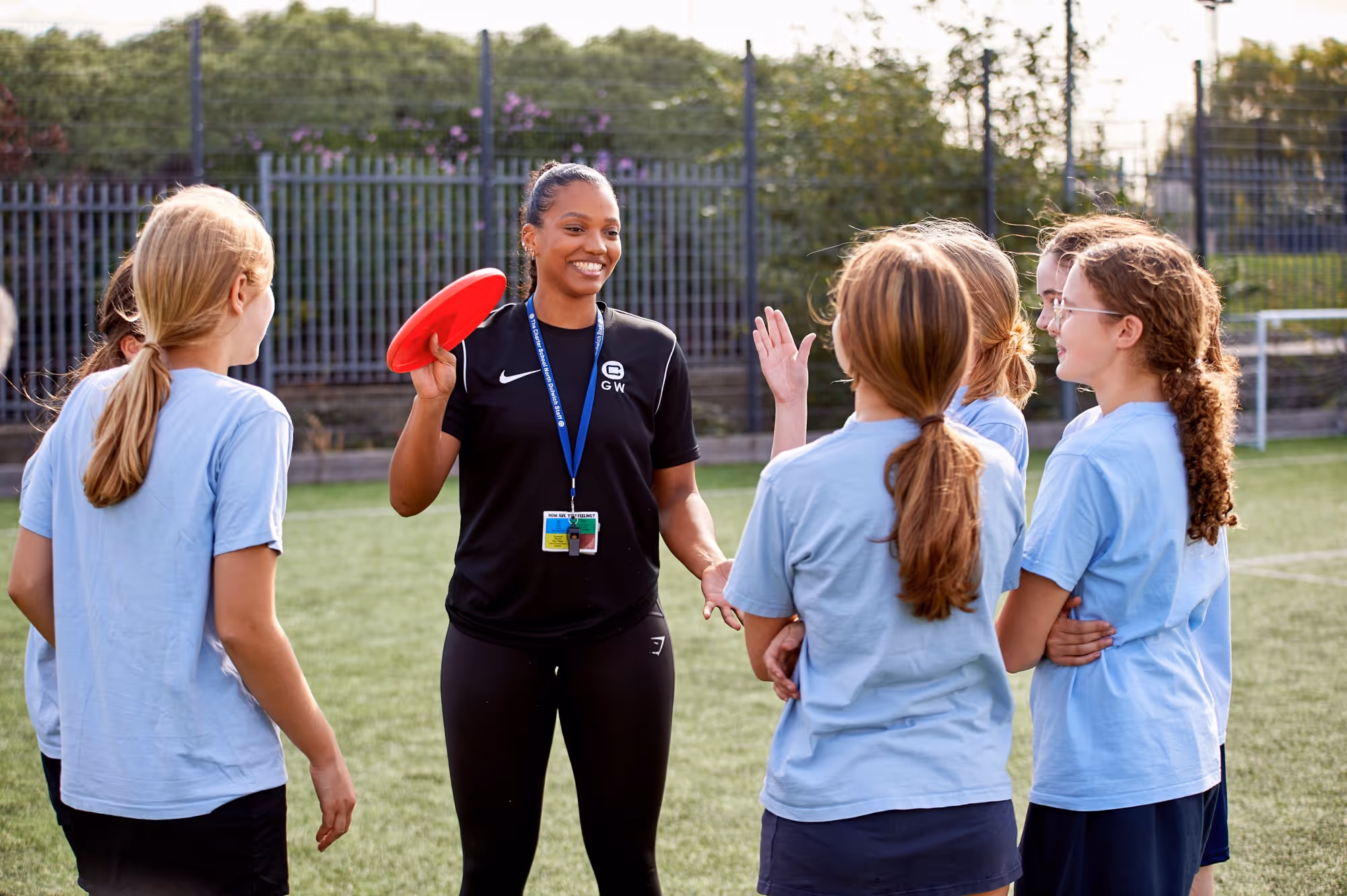 A female coach holding a red frisbee talks and smiles to a group of five girls in light blue shirts standing on a grassy sports field.