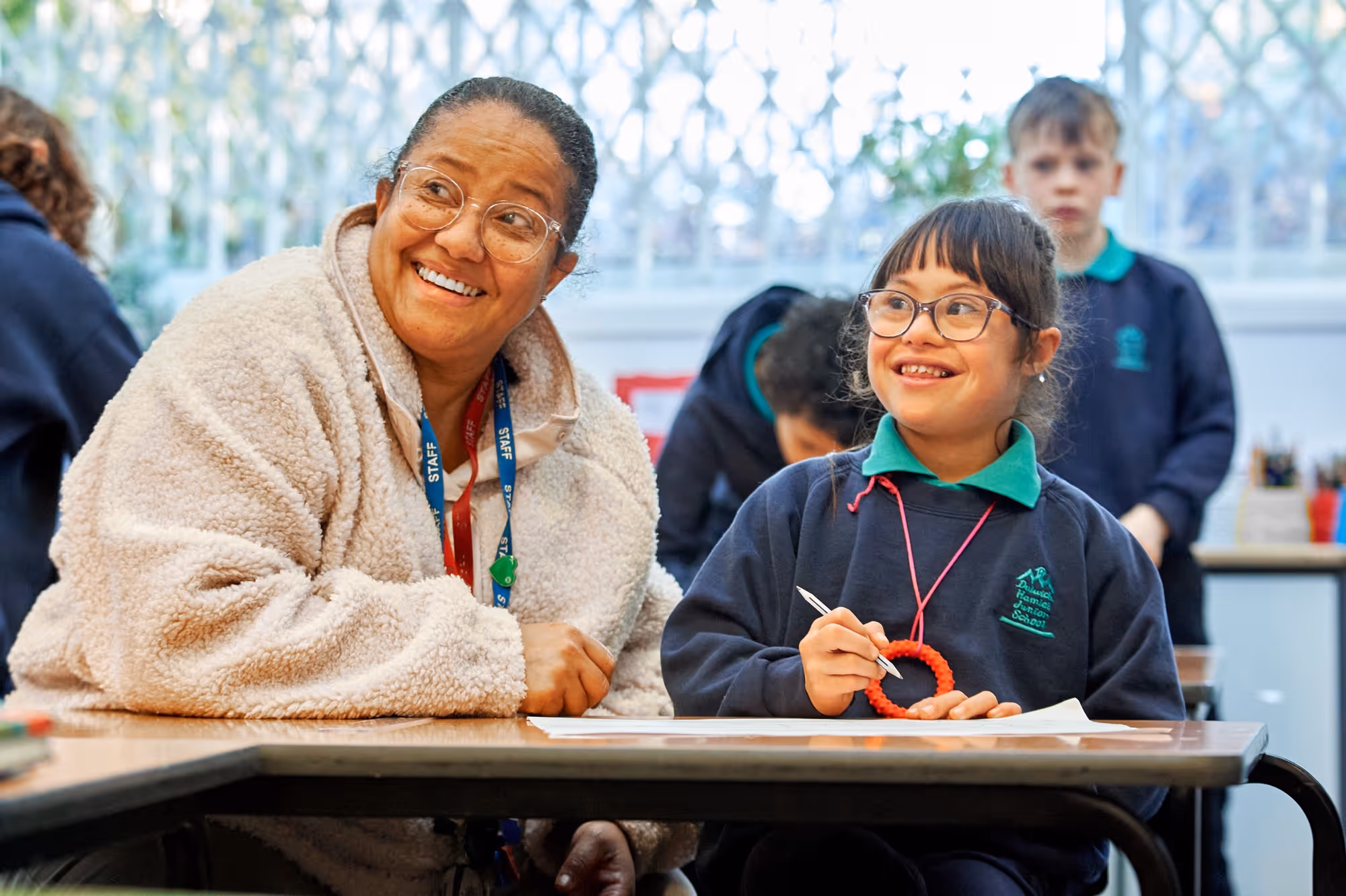 Smiling teacher wearing glasses and a fluffy jacket sits next to a smiling student with glasses and a green-collared school uniform at a classroom desk, both looking content.