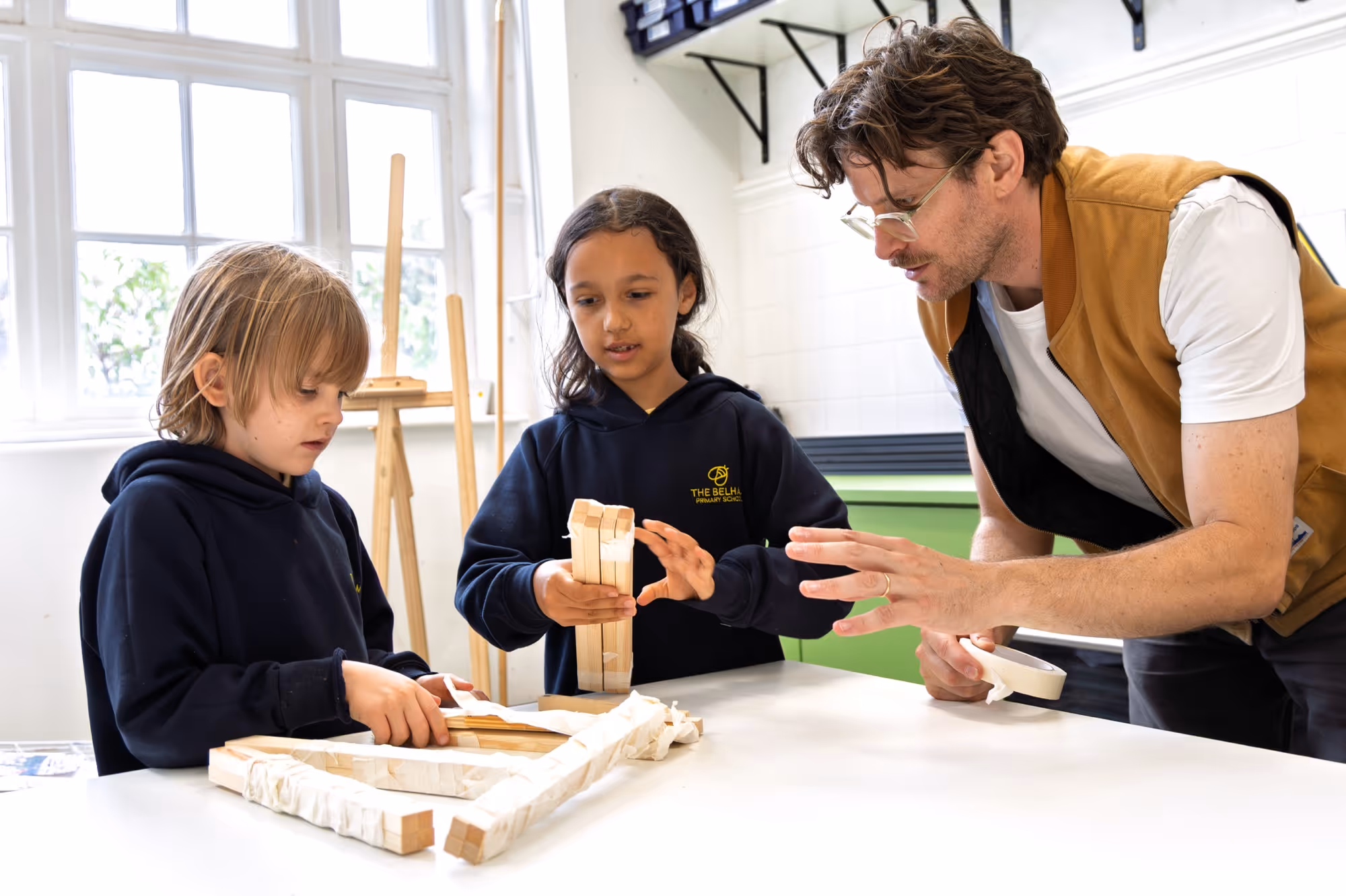 Two children in navy hoodies working with wooden blocks on a table while an adult man with glasses and a brown vest is guiding them in a bright room with large windows.