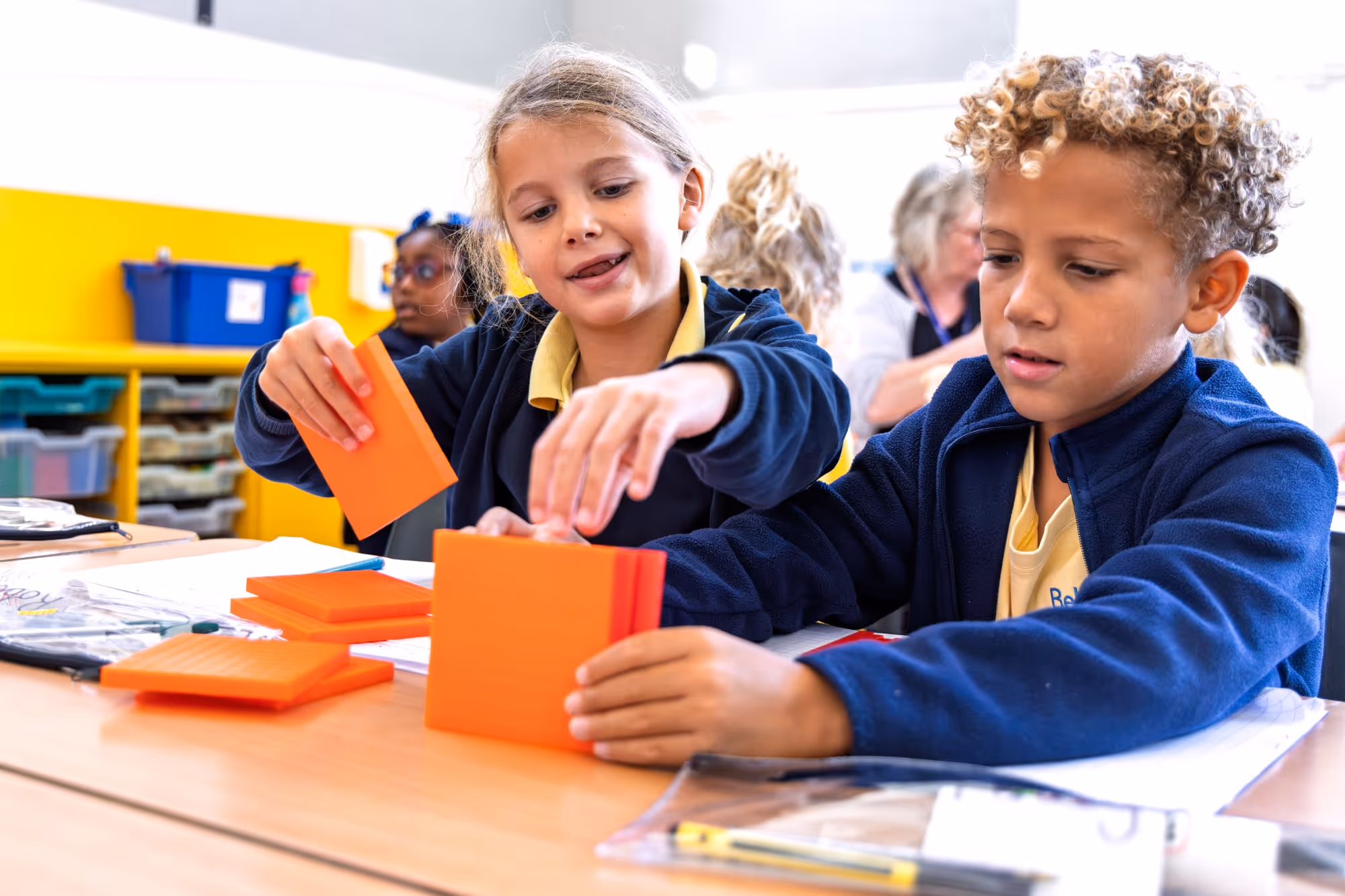 Two children in school uniforms sitting at a table and engaged in a hands-on activity with orange cards or blocks, with a classroom setting in the background.