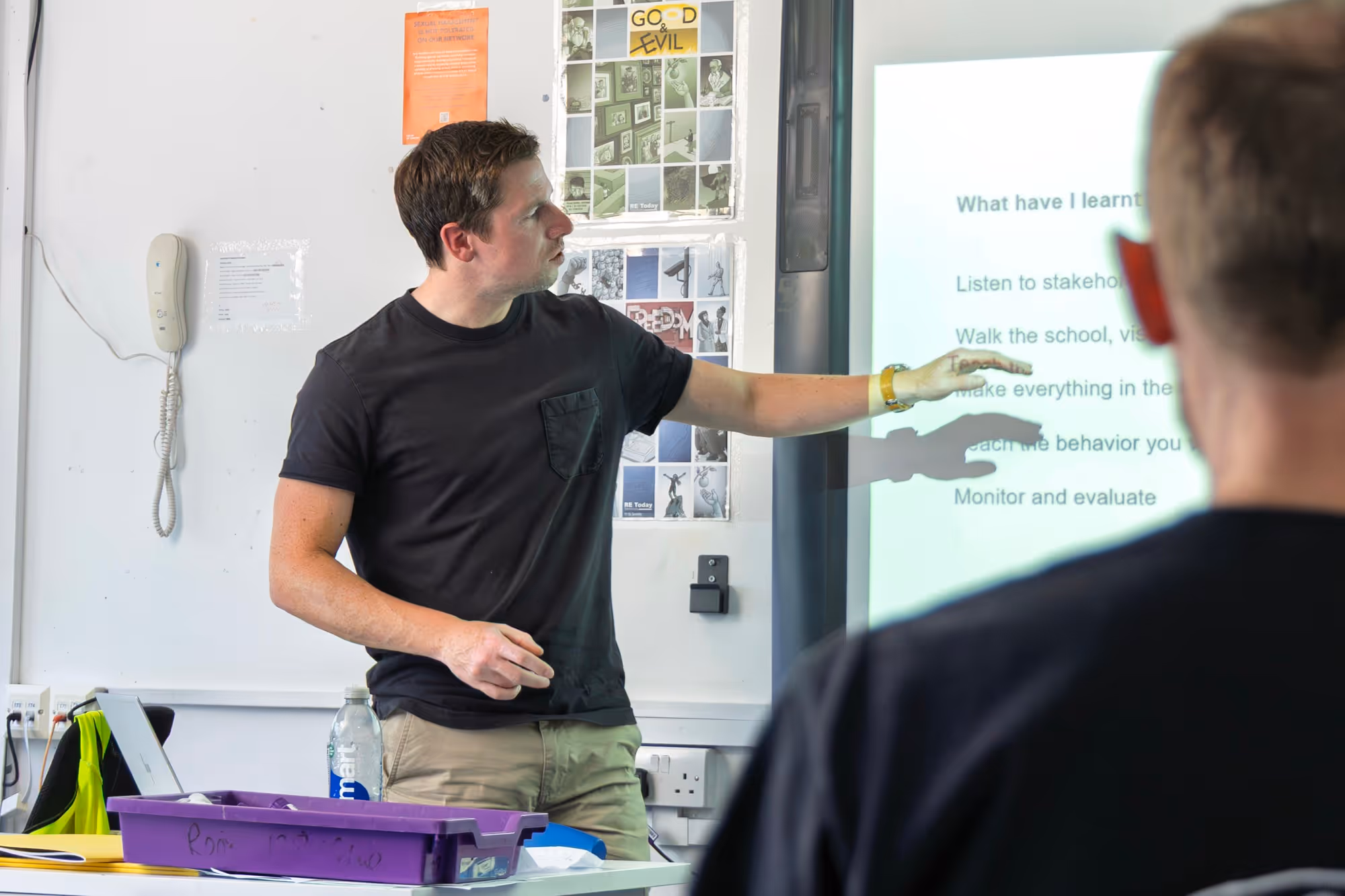 A man in a black t-shirt pointing at a projected slide during a presentation in a classroom, with another person in the foreground watching.